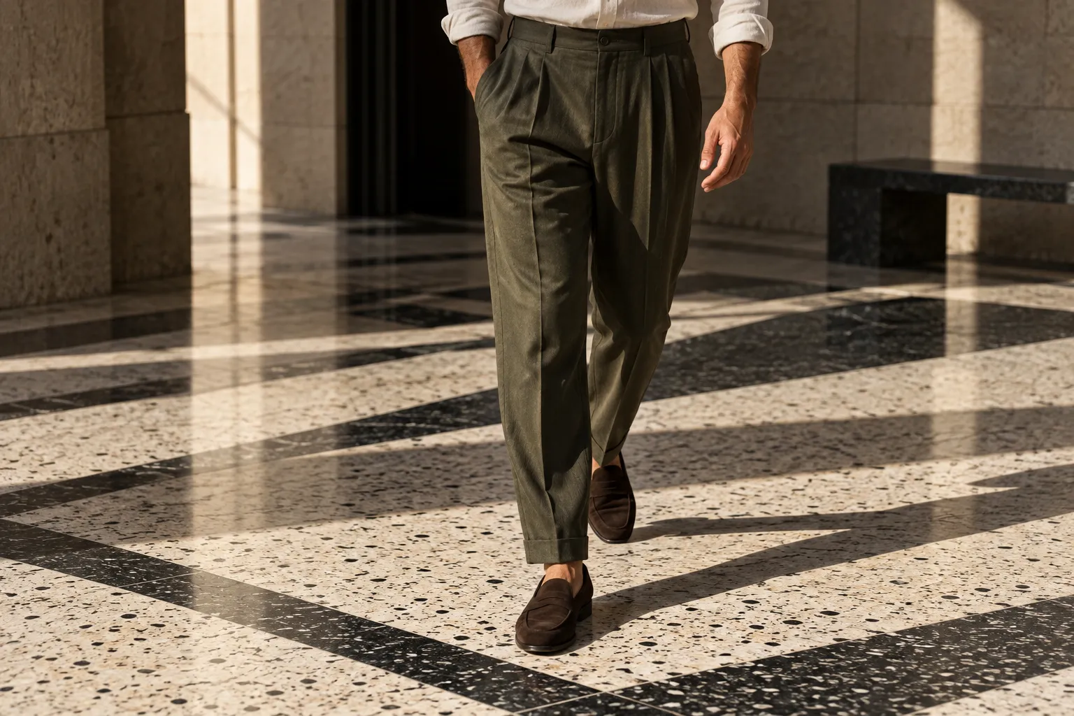 Deep olive high-rise pleated trousers and brown loafers captured mid-step across a geometric terrazzo floor